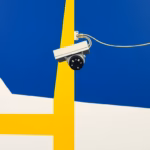 Suprematist abstract painting by Kazimir Malevich featuring a modern outdoor surveillance camera and electrical power cables. Minimalist composition of bold navy blue and vibrant yellow geometric shapes on a stark white background. Flat textures, sharp edges, avant-garde electrical concept, clean geometric abstraction. No humans, no text overlay.