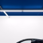 Suprematist abstract painting in the style of Kazimir Malevich depicting a garage ceiling fitted with linear LED batten lights glowing brightly above a car silhouette. Minimalist composition of bold navy blue and vibrant yellow geometric shapes on a stark white background. Flat textures, sharp edges, avant-garde electrical concept, no humans, no text overlay.