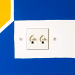 Suprematist abstract painting inspired by Kazimir Malevich featuring a recessed French wall electrical socket with two adjustable metal claw fixing plates gripping a sealed plaster back box. Minimalist composition of bold navy blue and vibrant yellow geometric shapes on a stark white background. Flat textures, sharp clean edges, avant-garde electrical renovation concept, modern editorial illustration style. No humans, no text, no lettering, no watermark.