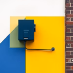 Suprematist abstract painting by Kazimir Malevich featuring a wall-mounted outdoor alarm siren with a strobe flash on a brick facade. Minimalist composition of bold navy blue and vibrant yellow geometric shapes on a stark white background. Flat textures, sharp edges, avant-garde electrical concept. No humans, no text.